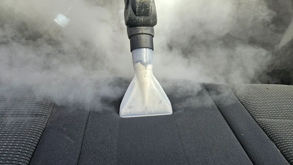 Duză transparentă de curățare poziționată pe materialul textil închis la culoare al unui scaun auto.