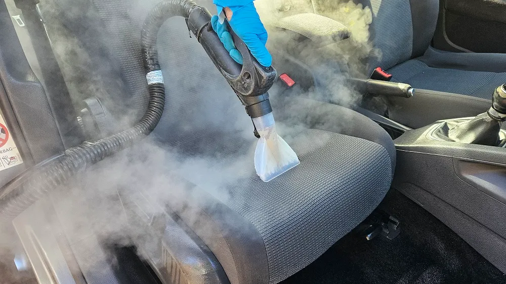 Operator cu mănușă albastră deplasează accesoriul de curățare pe șezutul unui scaun auto gri.